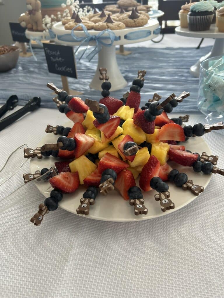Plate of fruit kabobs arranged in a circle, each skewer topped with a small bear decoration and filled with blueberries, strawberries, and pineapple, with cookies and cupcakes visible in the background.