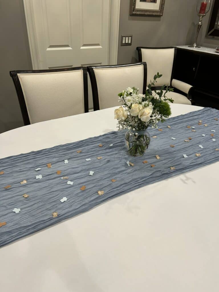 Dining table with a white tablecloth, blue crinkled table runner, gold and blue bear‑and‑cloud confetti, and a floral centerpiece of white roses, baby’s breath, and greenery.