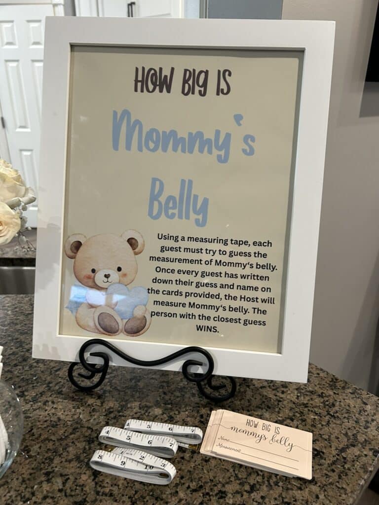 Framed “How Big Is Mommy’s Belly” baby shower game sign displayed with measuring tapes and matching guess cards on a countertop.