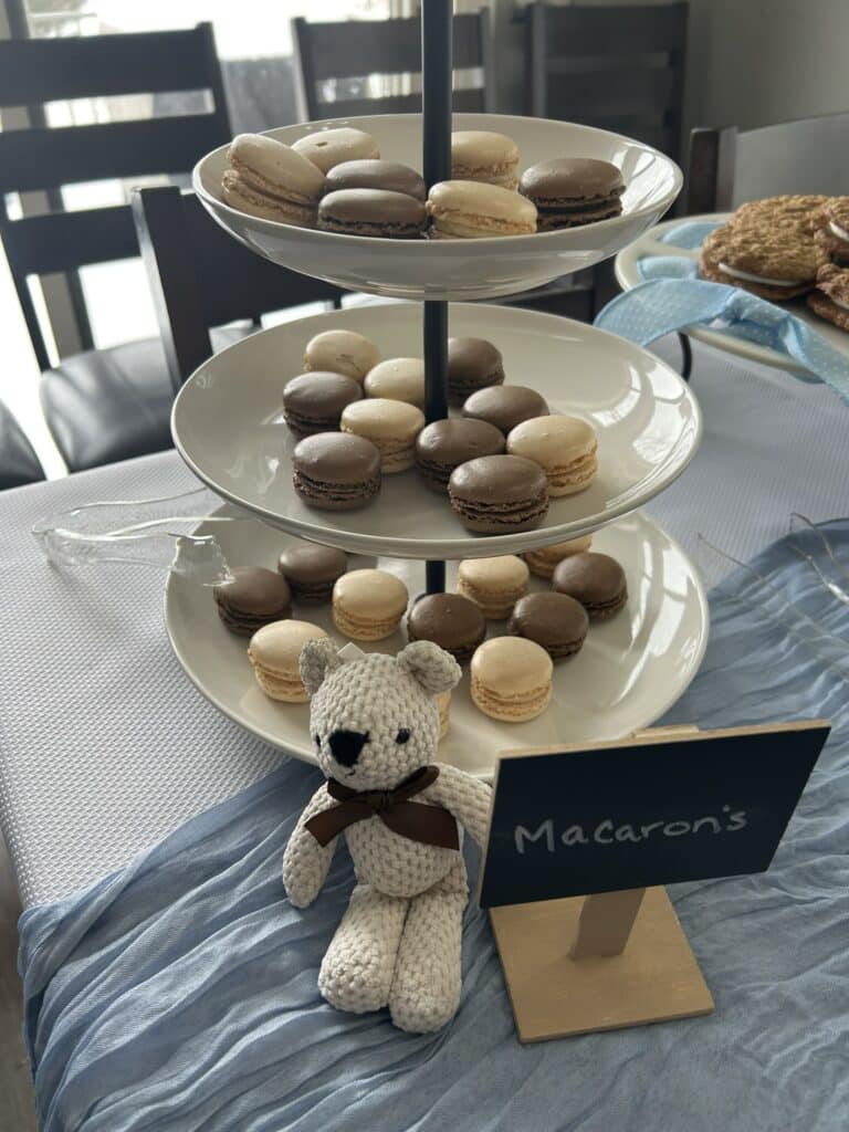 Three‑tiered stand filled with vanilla and chocolate macarons in neutral tones, displayed with a small crocheted teddy bear and a chalkboard sign on a dessert table.
