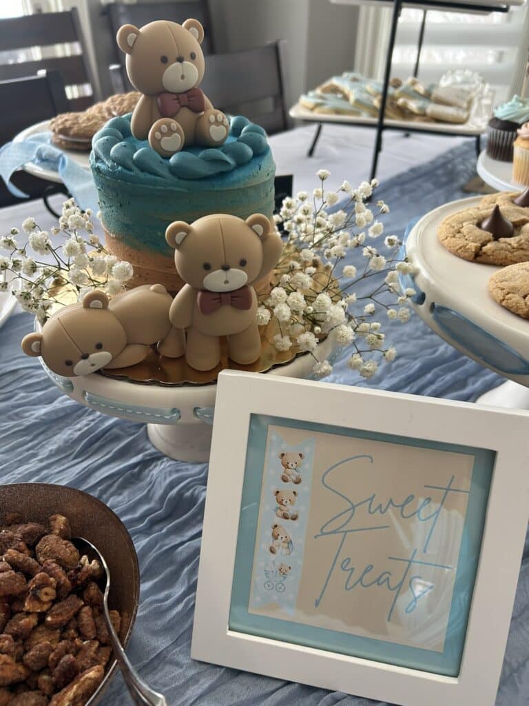 Framed “Sweet Treats” sign with teddy bear illustrations displayed in front of a bear‑topped cake, cookies, and a bowl of candied nuts on a blue‑covered dessert table.