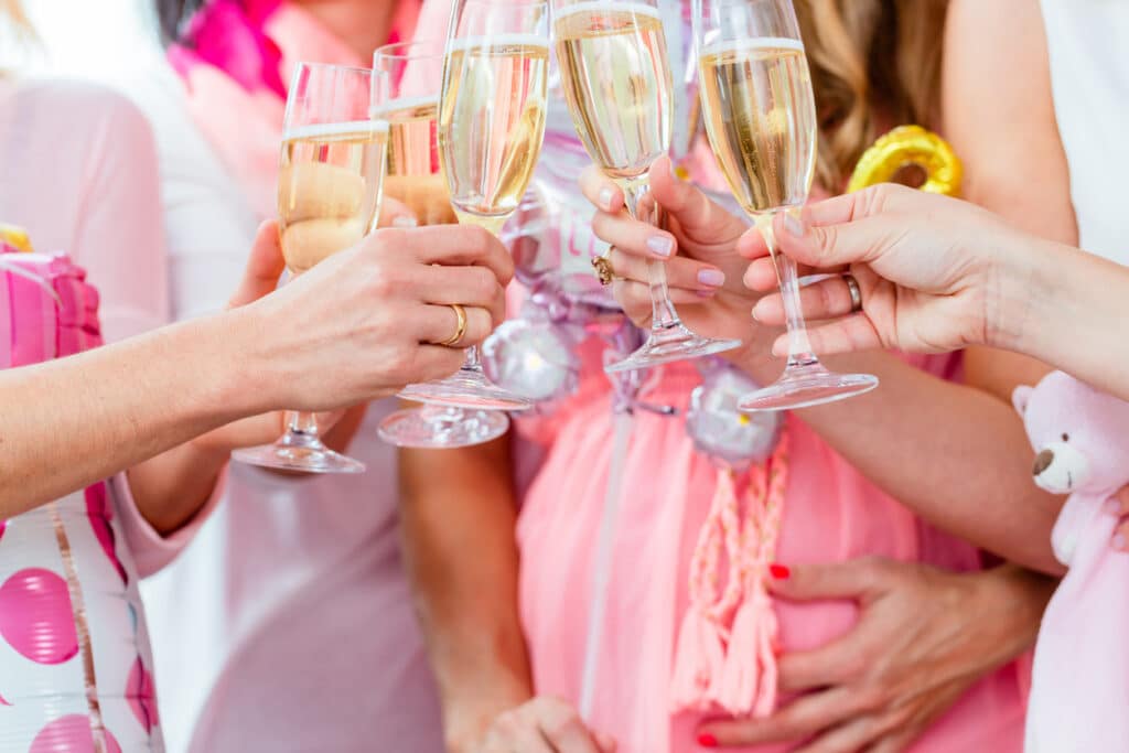 Group of people raising drink glasses in a celebratory toast, with pink accents, a stuffed bear, and a hand resting on a pregnant belly.