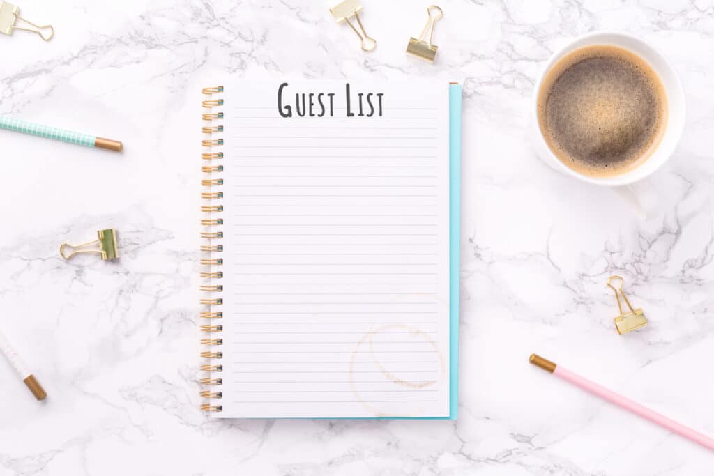 Flat lay of a notebook labeled “Guest List” with pens, coffee, and office supplies, illustrating how to plan a baby shower by organizing who to invite.