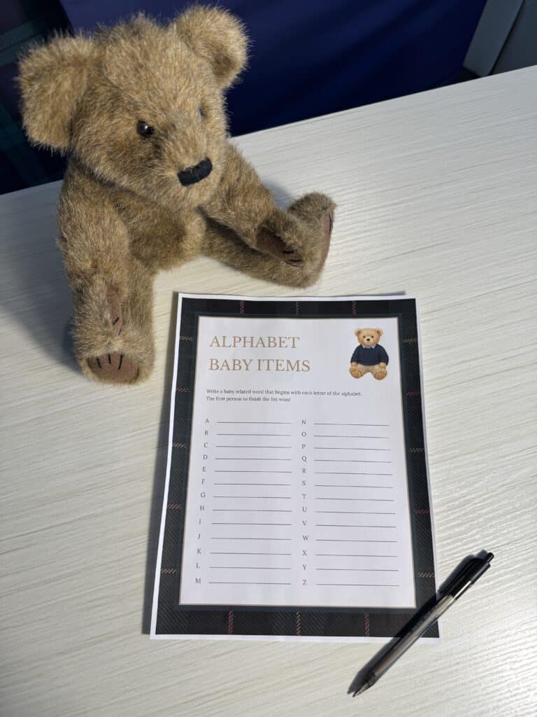 Teddy bear sitting beside an “Alphabet Baby Items” baby shower game card and a black pen on a light wooden table, featuring A–Z lines for guests to list baby‑related words.