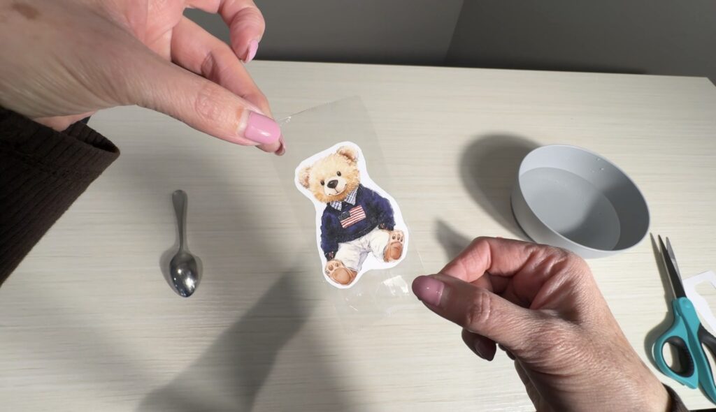 Hands holding a cut‑out teddy bear illustration covered with clear tape during the tape transfer craft process.