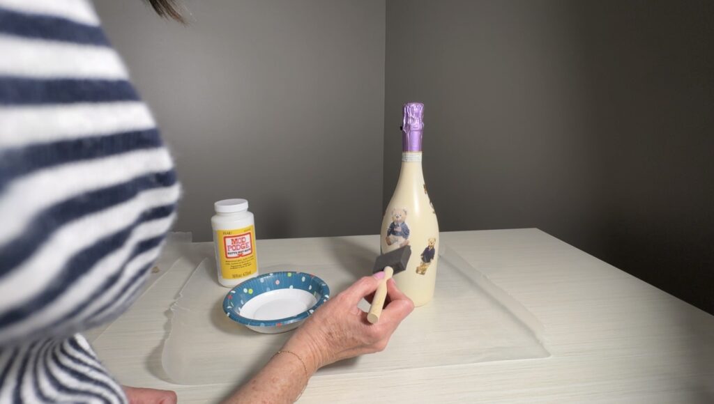 Applying a thin layer of Mod Podge with a foam brush to seal teddy‑bear decoupage on a painted wine bottle.