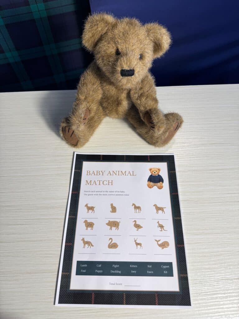 Teddy bear sitting beside a “Baby Animal Match” baby shower game card and a black pen on a light wooden table, featuring illustrated adult animals with blank spaces for guests to write the correct baby animal names.
