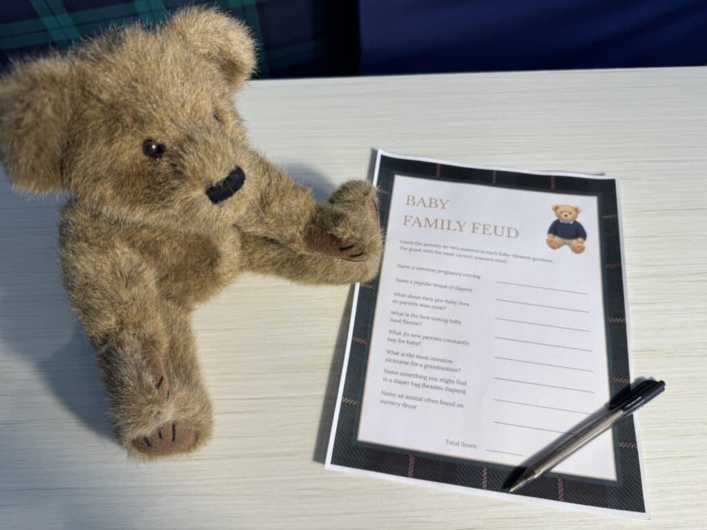 Teddy bear sitting beside a “Baby Family Feud” baby shower game card and a black pen on a white wooden table, featuring baby‑themed survey questions with blank lines for answers.