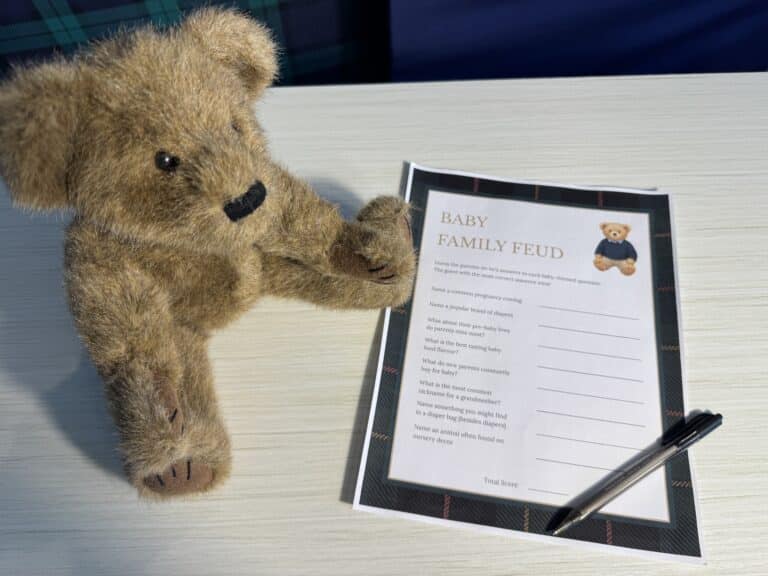 Teddy bear sitting beside a “Baby Family Feud” baby shower game card and a black pen on a white wooden table, featuring baby‑themed survey questions with blank lines for answers.