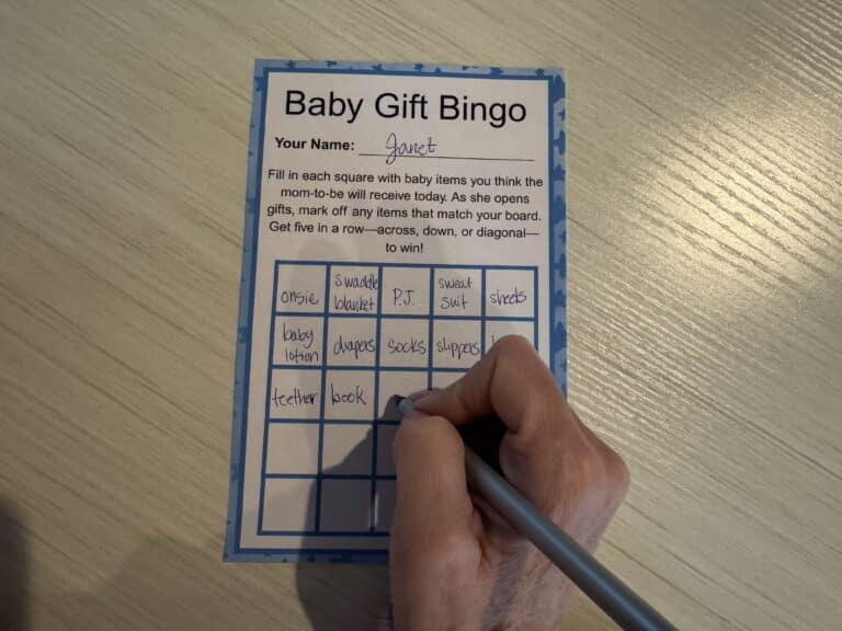 Hand filling out a Baby Gift Bingo card on a wooden table, with squares labeled for common baby shower gifts such as diapers, onesies, books, and swaddles.