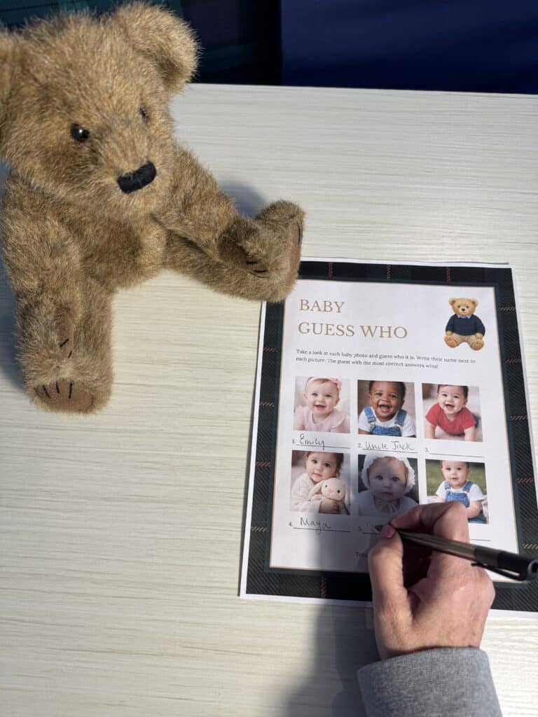 Hand holding a pen while filling out a “Baby Guess Who” baby shower game card on a light wooden table, with six baby photos and a teddy bear beside the sheet.