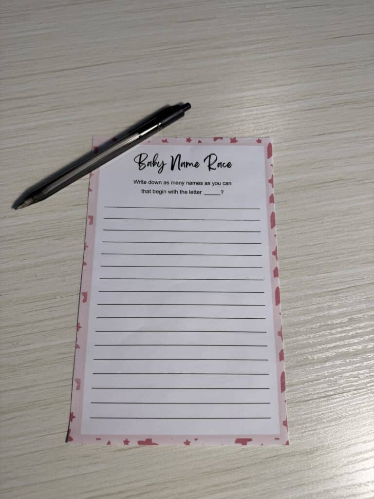 “Baby Name Race” baby shower game card with a pink border and a black pen resting on top, placed on a light wooden table, featuring blank lines for guests to write baby names starting with a chosen letter.
