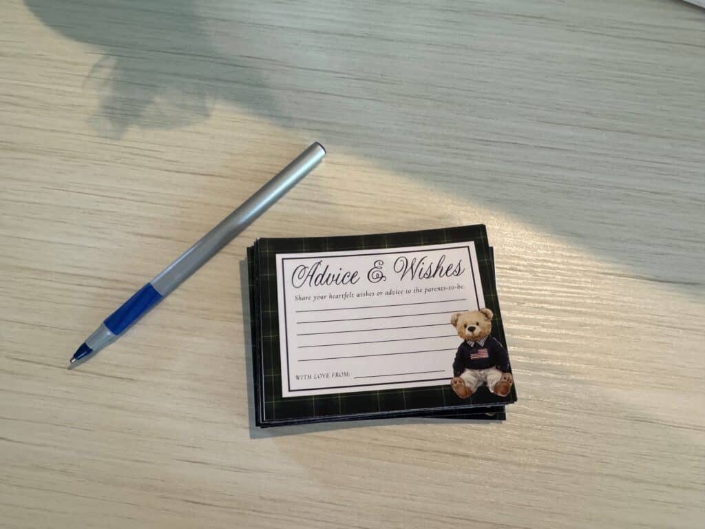 Close-up of baby shower advice cards featuring a classic teddy bear in a navy sweater, displayed on a light wood surface with a blue pen.