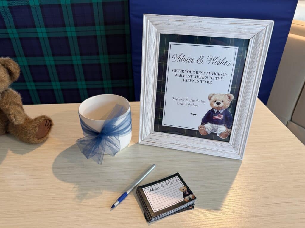 Baby shower advice cards displayed on a table with a framed sign, navy ribbon box, and classic teddy bear in a Ralph Lauren–inspired theme.