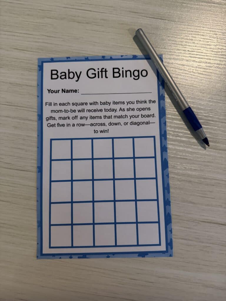 Blank Baby Gift Bingo card with a blue border and baby‑themed icons, displayed on a wooden table with a blue pen beside it.
