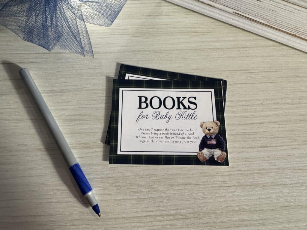 Close‑up of a blue plaid “Books for Baby” card with a teddy bear illustration, placed on a wooden surface beside a pen and blue tulle.