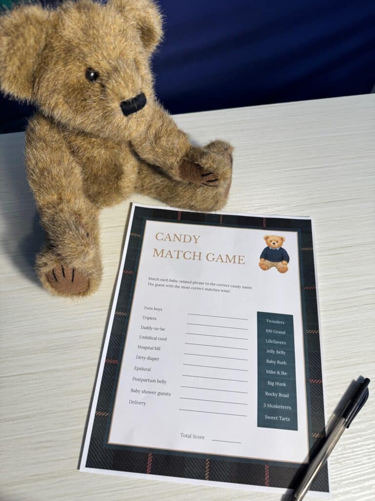 Teddy bear sitting beside a “Candy Match Game” baby shower game card and a black pen on a light wooden table, featuring candy names and baby‑related phrases for guests to match.