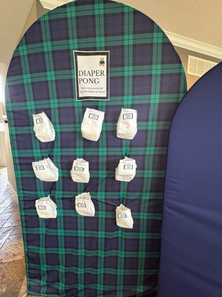 Plaid‑backdrop Diaper Pong baby shower game display with nine diapers labeled with point values from 10 to 50 and a sign that reads “Diaper Pong — Toss a ping pong ball into the diaper to score.”