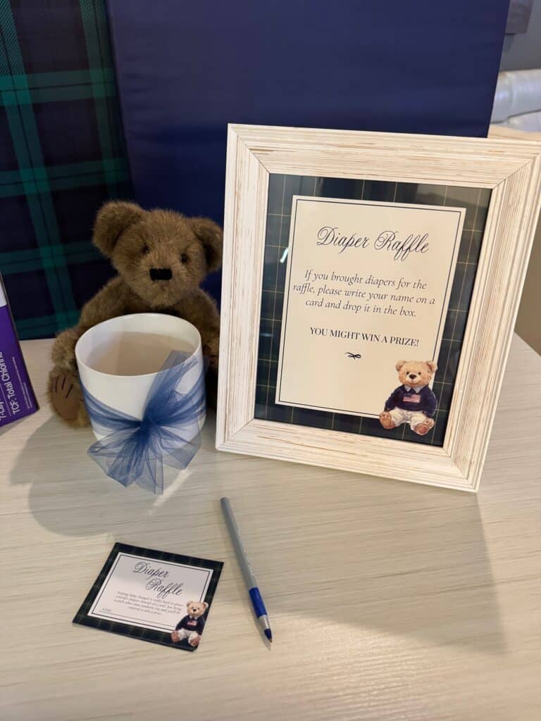 Baby shower diaper raffle station with a framed sign, teddy bear, blue‑ribbon cup, pen, and matching raffle cards displayed on a plaid‑covered tabletop.