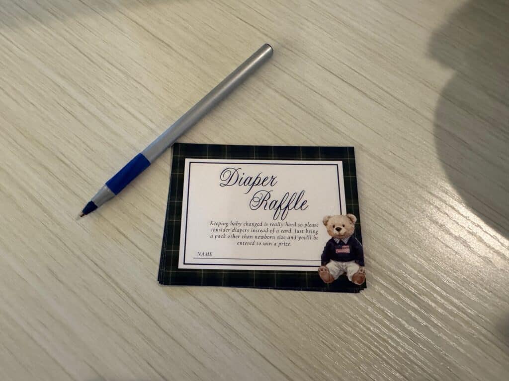 Close‑up of a plaid diaper raffle card with a teddy bear illustration, displayed on a wooden surface beside a blue and silver pen.