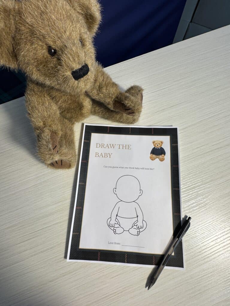 Teddy bear sitting beside a “Draw the Baby” baby shower game card on a white table, with a black pen next to the sheet featuring a blank baby outline for guests to draw the baby’s face.
