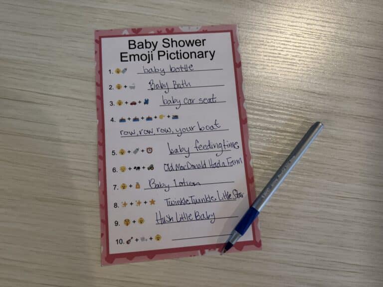 Baby Shower Emoji Pictionary game sheet with a pink border, handwritten answers, and a blue pen displayed on a wooden table.