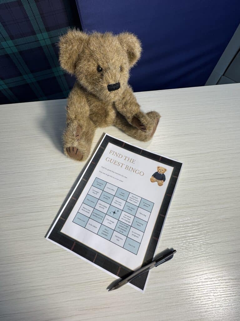 Teddy bear sitting beside a “Find the Guest Bingo” baby shower game card and a black pen on a light wooden table, featuring a 5×5 grid of guest‑prompt squares.
