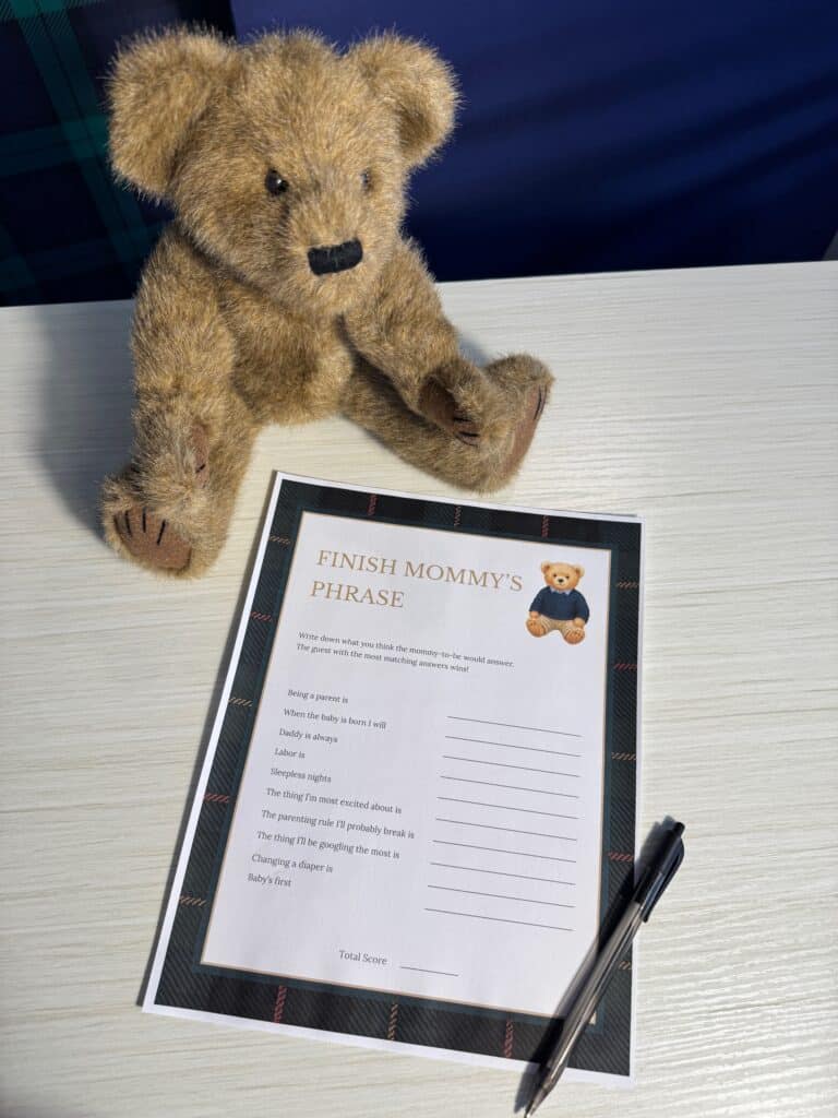 Teddy bear sitting beside a “Finish Mommy’s Phrase” baby shower game card and a black pen on a white wooden table, featuring fill‑in‑the‑blank prompts for guests to guess how the mom‑to‑be would complete each phrase.