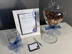 Baby shower “Guess How Many” game setup with a jar filled with Teddy Grahams, a framed instruction sign, guess cards, and a small container with a pen.