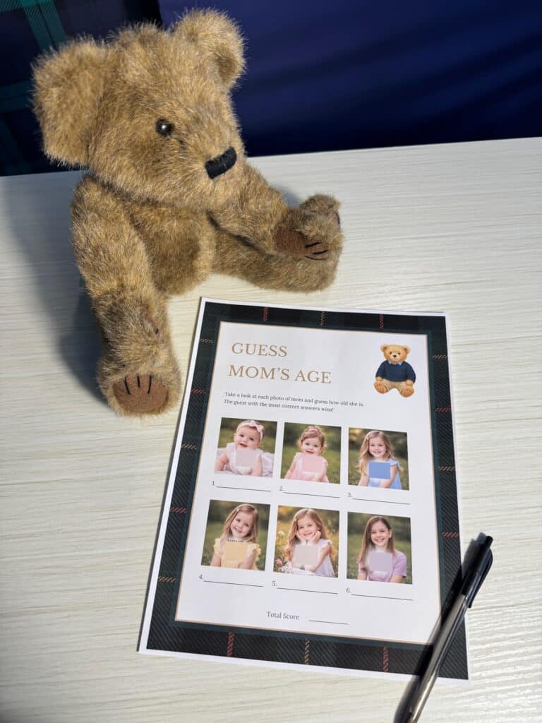 Teddy bear sitting beside a “Guess Mom’s Age” baby shower game card and a black pen on a white wooden table, featuring six childhood photos with blank lines for guests to guess her age.