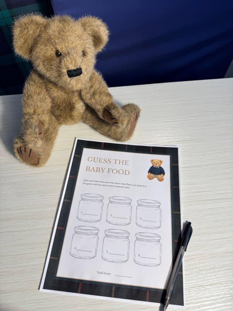 Teddy bear sitting beside a “Guess the Baby Food” baby shower game card and a black pen on a light wooden table, featuring six numbered jars with blank lines for guests to guess each baby food flavor.