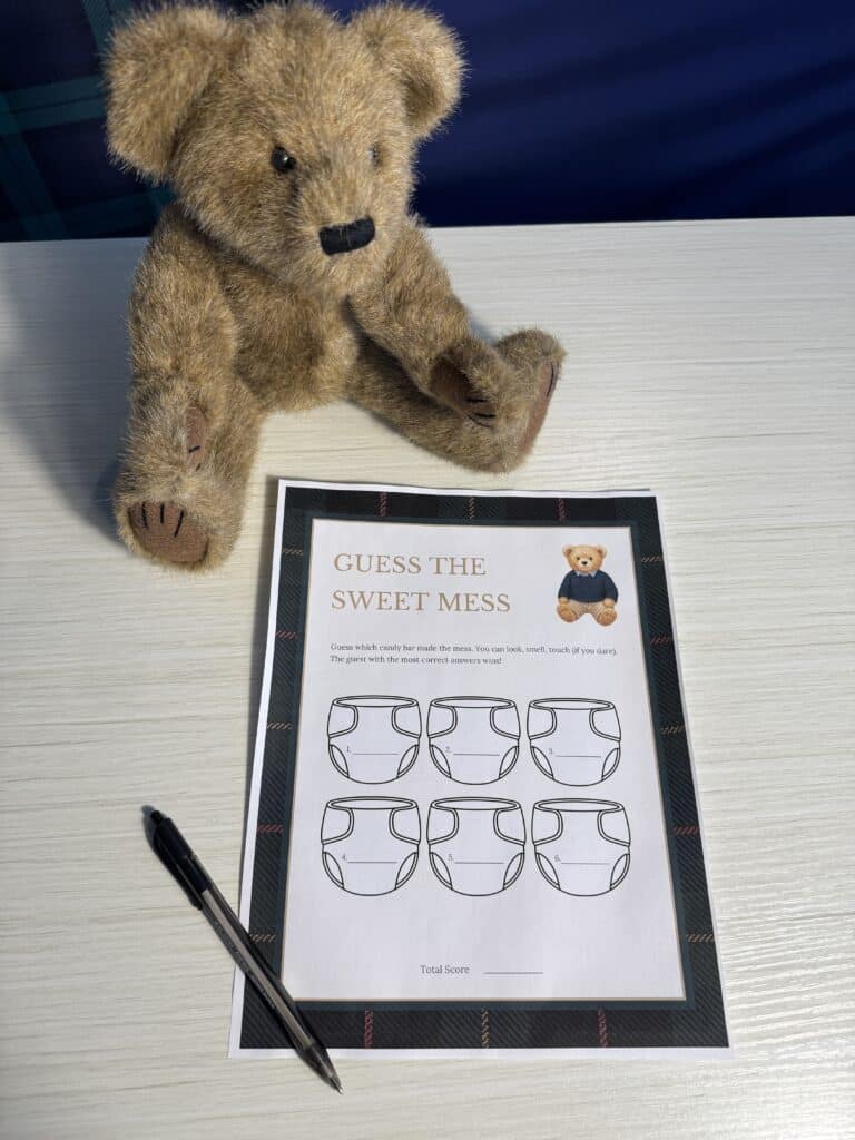 Teddy bear sitting beside a “Guess the Sweet Mess” baby shower game card and a black pen on a white wooden table, featuring six labeled jars (A–F) with blank lines for guests to guess the candy bar.