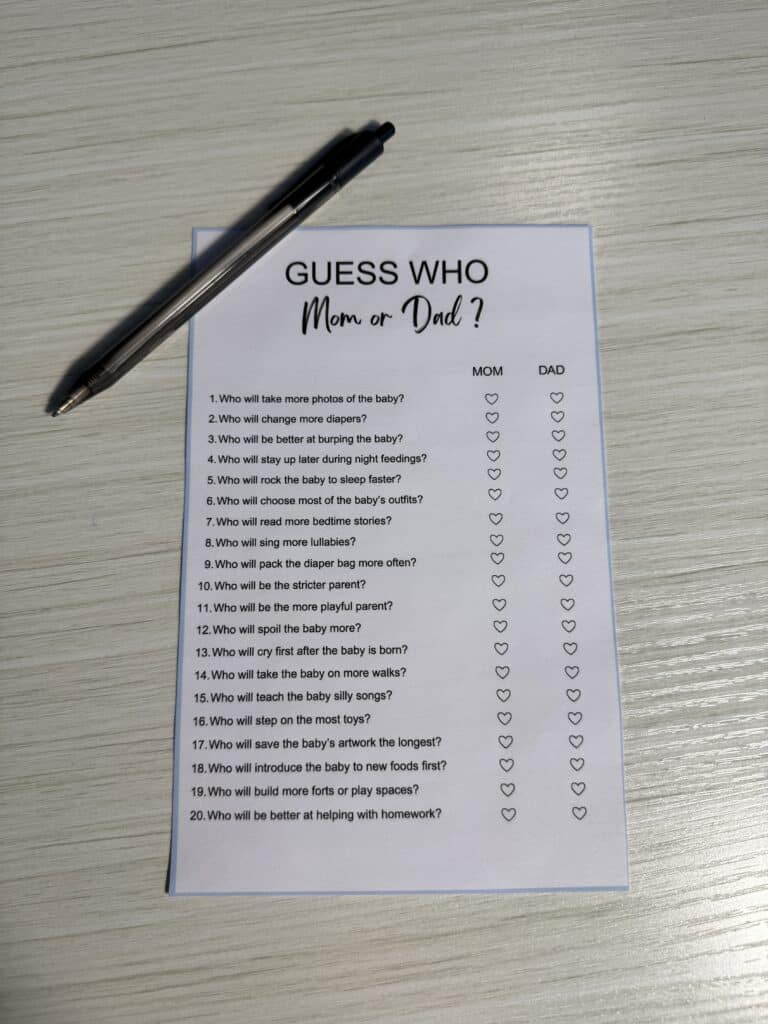 “Guess Who: Mom or Dad?” baby shower game card on a light wooden table with a black pen beside it, featuring parenting‑themed questions and heart icons for marking answers.