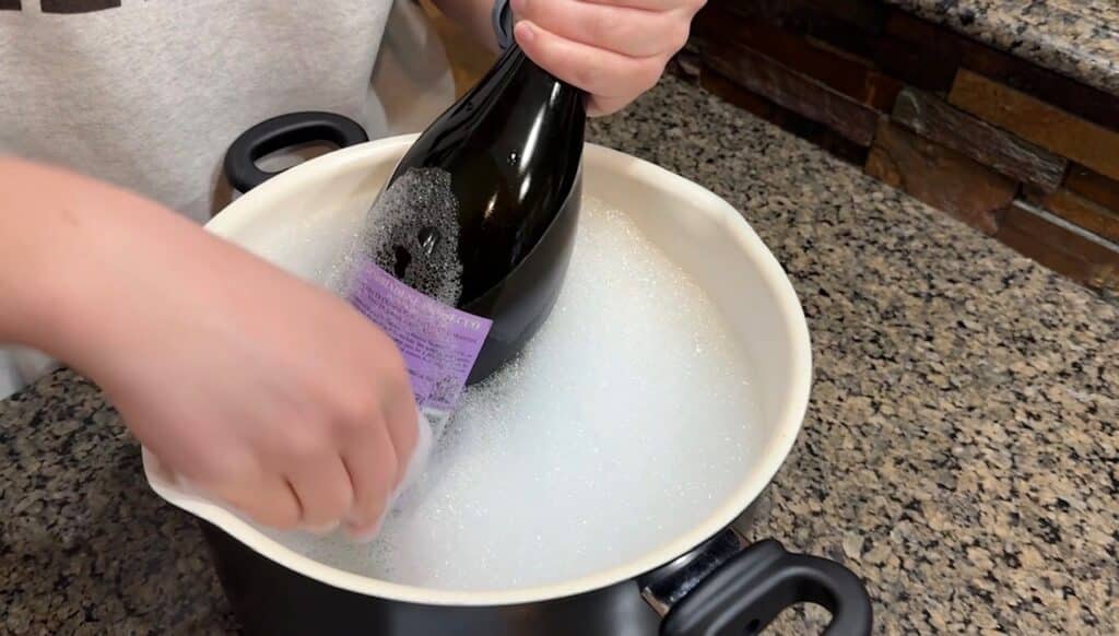 Person cleaning a wine bottle in warm soapy water as part of a tutorial on how to remove wine bottle labels for DIY projects.