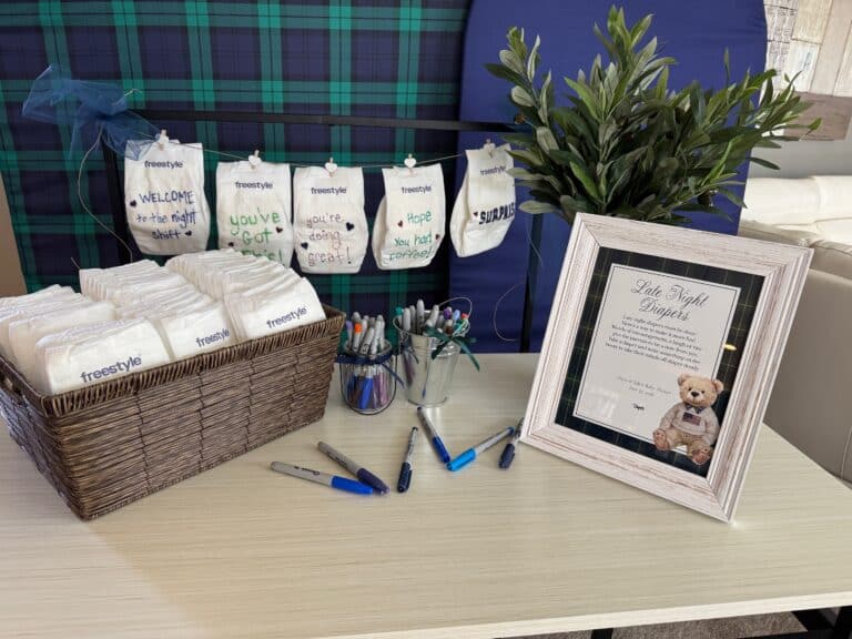 Baby shower table with a basket of blank diapers for guests to write late-night diaper notes, with sample decorated diapers hanging on a mini clothesline behind the table.
