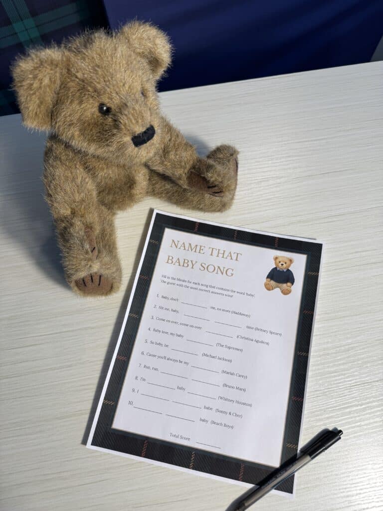 Teddy bear sitting beside a “Name That Baby Song” baby shower game card and a black pen on a light wooden table, featuring a list of fill‑in‑the‑blank song titles that use the word “baby.”