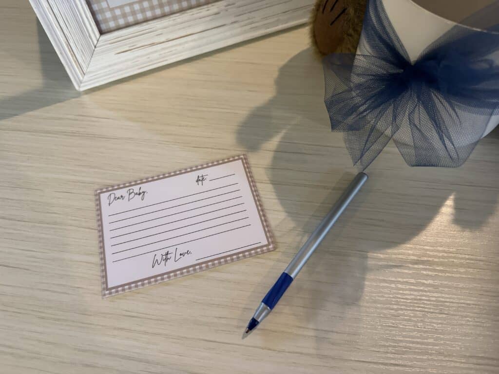 Close‑up of a beige gingham “Dear Baby” printable note card with writing lines, a pen, and a blue tulle bow decoration on a tabletop.