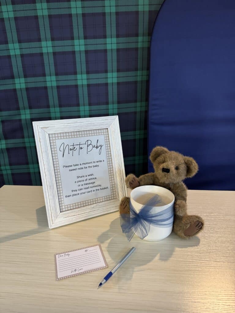 Close‑up of a baby shower “Note to Baby” sign in a white frame, displayed with a small teddy bear, a blue‑ribbon cup, a pen, and a matching note card on a tabletop.