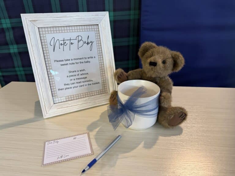 Baby shower “Note to Baby” station with framed sign, teddy bear, blue ribbon cup, pen, and matching note card on a light wooden table.