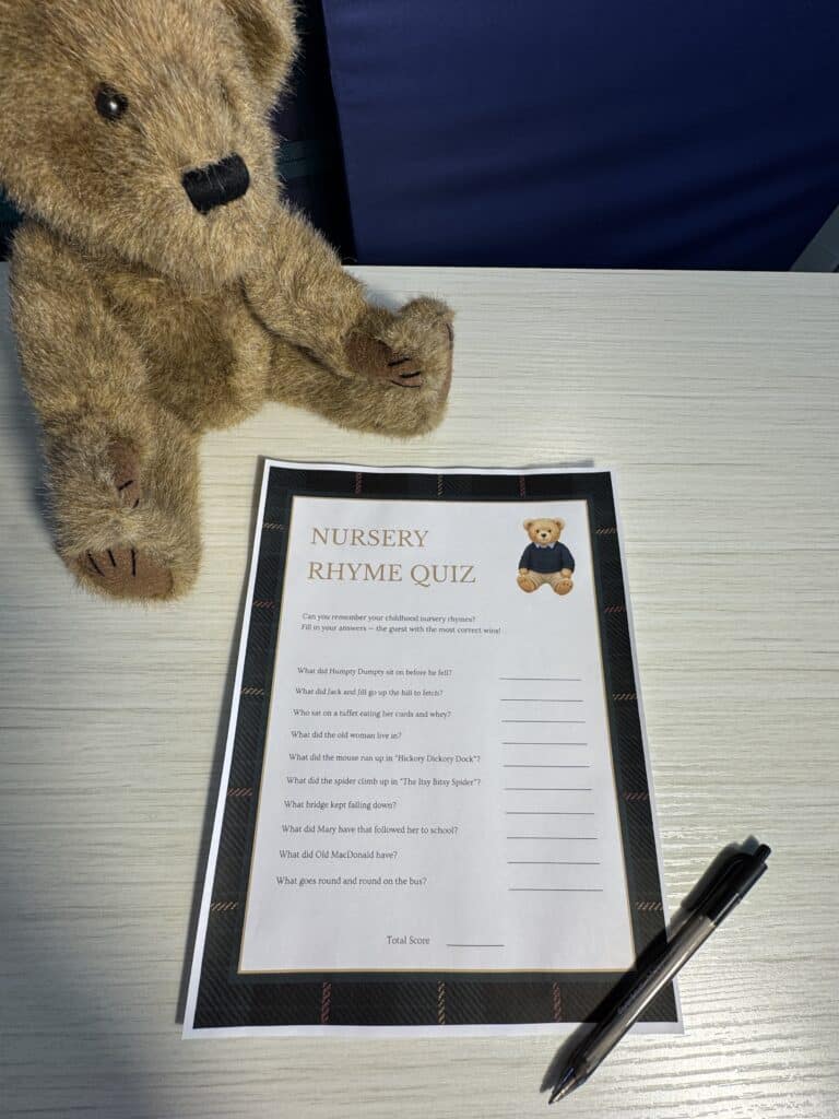 Teddy bear sitting beside a “Nursery Rhyme Quiz” baby shower game card and a black pen on a light wooden table, featuring classic nursery‑rhyme questions with blank lines for answers.