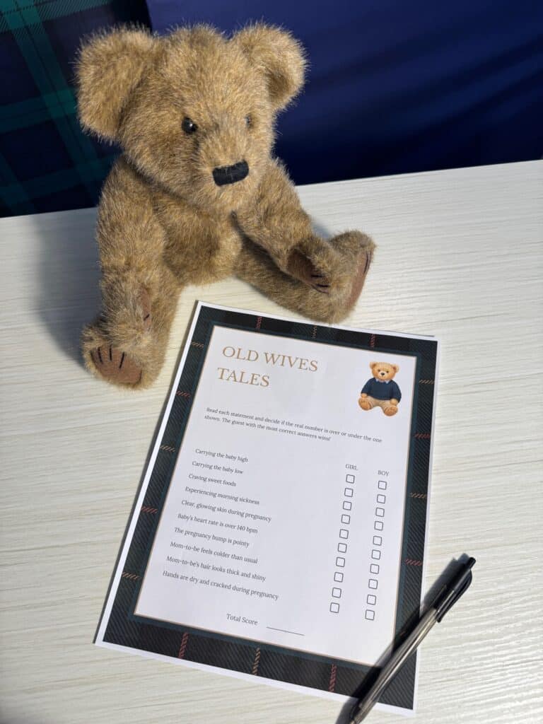 Teddy bear sitting beside an “Old Wives Tales” baby shower game card and a black pen on a white wooden table, featuring boy‑versus‑girl prediction prompts with checkboxes.
