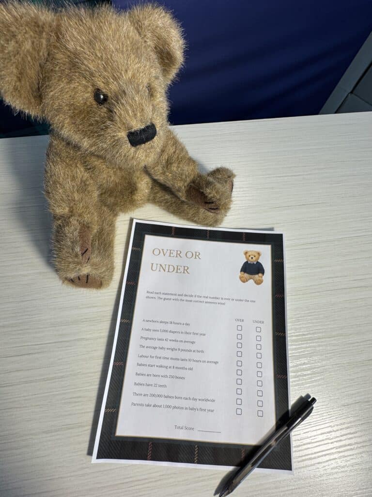 Teddy bear sitting beside a “Nursery Rhyme Quiz” baby shower game card and a black pen on a light wooden table, featuring classic nursery‑rhyme questions with blank lines for answers.