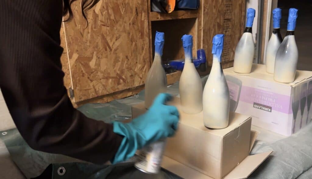 Person spray‑painting wine bottles white, with the tops taped off for protection.