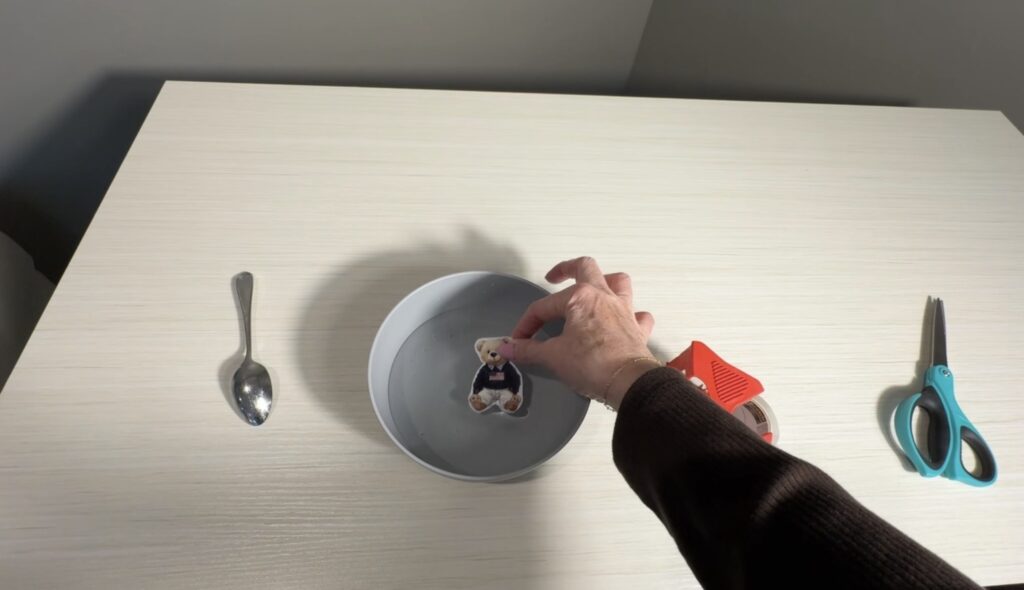 Hand placing a taped teddy bear image into a bowl of water during the tape transfer process.