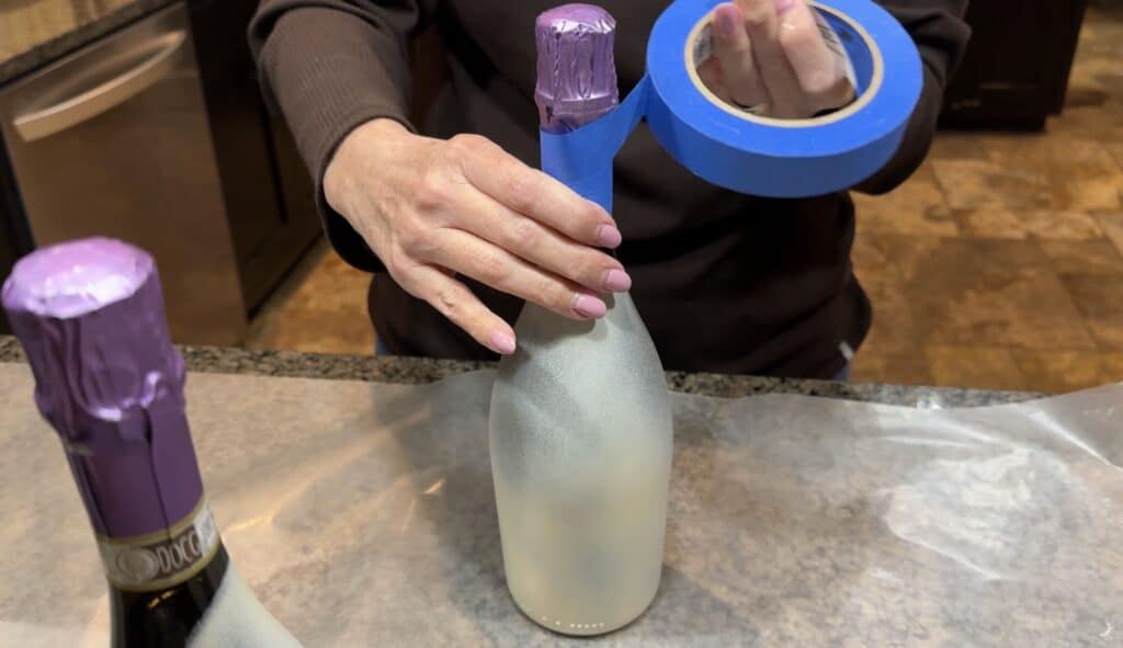 Person applying blue painter’s tape around the neck of a painted wine bottle to protect the top before decorating.