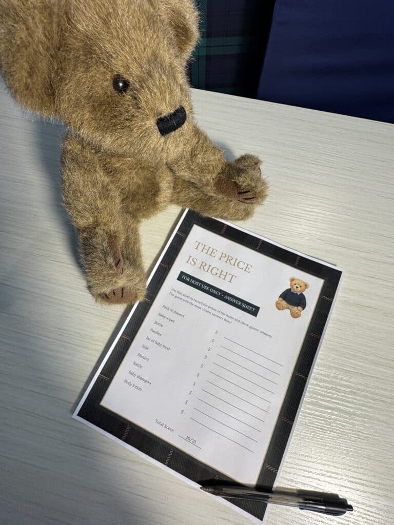 Teddy bear sitting beside a “The Price Is Right” baby shower game card and a black pen on a light wooden table, featuring a list of baby‑item prices for guests to guess.