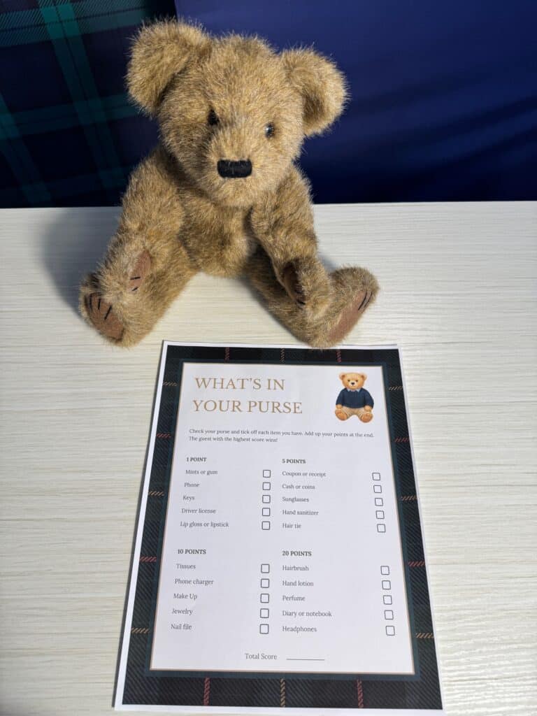 Teddy bear sitting beside a “What’s in Your Purse” baby shower game card and a black pen on a light wooden table, featuring a points‑based checklist of common purse items.