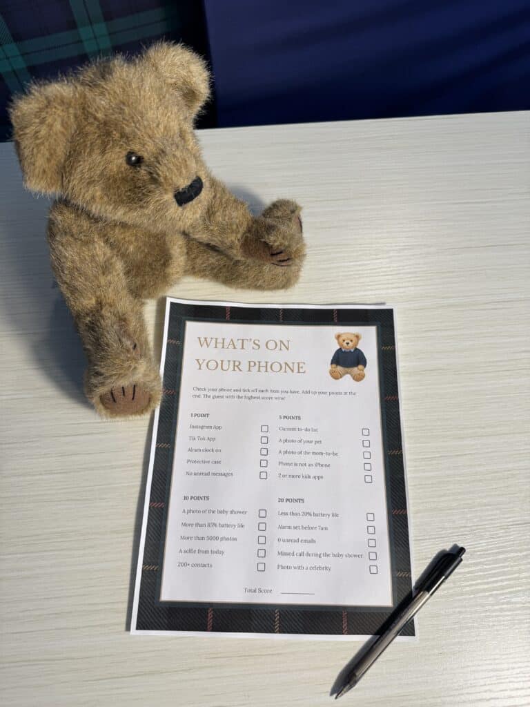 Teddy bear sitting beside a “What’s On Your Phone” baby shower game card and a black pen on a light wooden table, featuring a checklist of phone items with different point values.