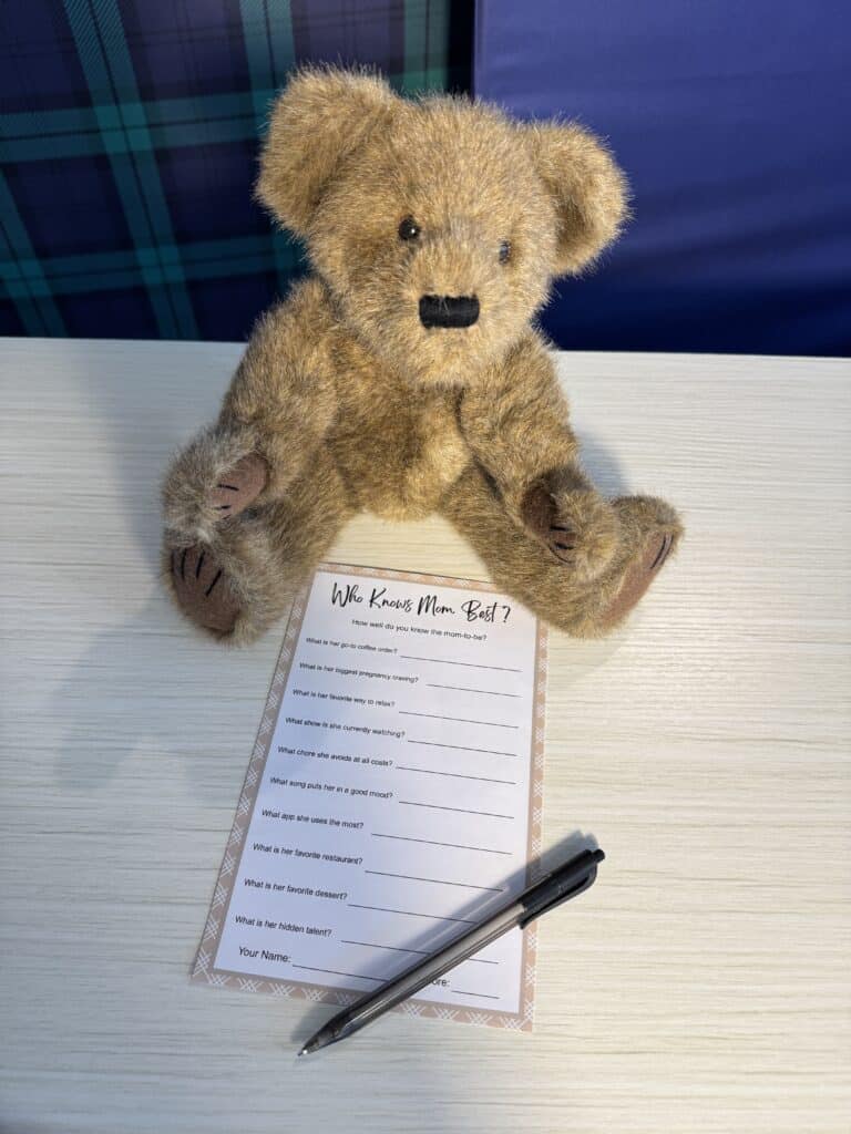 “Guess Who: Mom or Dad?” baby shower game card on a light wooden table with a black pen beside it, featuring a list of parenting‑themed questions with heart icons for marking answers.