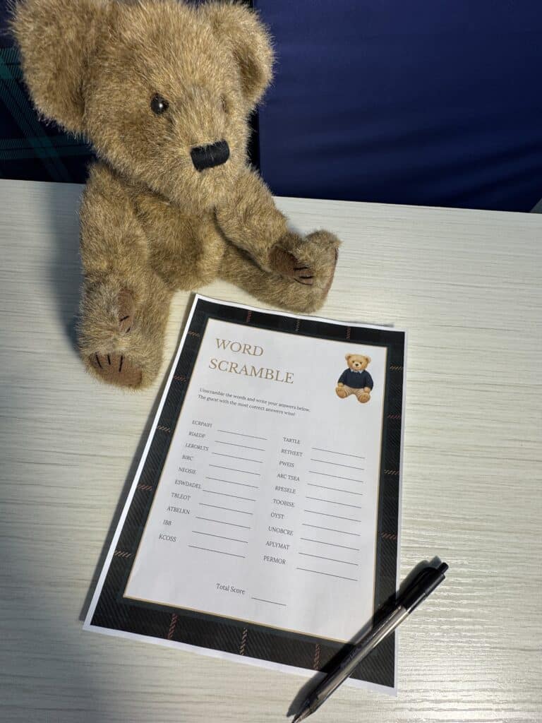 Teddy bear sitting beside a “Word Scramble” baby shower game card and a black pen on a light wooden table, featuring scrambled words and blank lines for guests to solve.